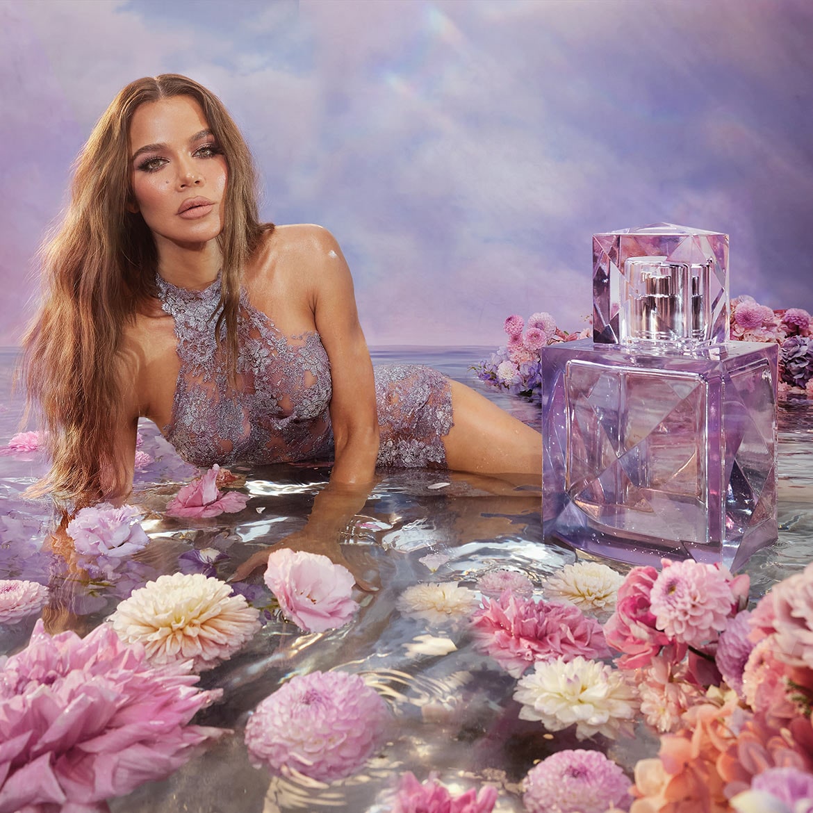 Na obrázku je Khloe Kardashian v levanduľových čipkovaných šatách, ležiaca vo vode s ružovými kvetmi, vedľa nej je fľaša parfumu v tvare kryštálu.