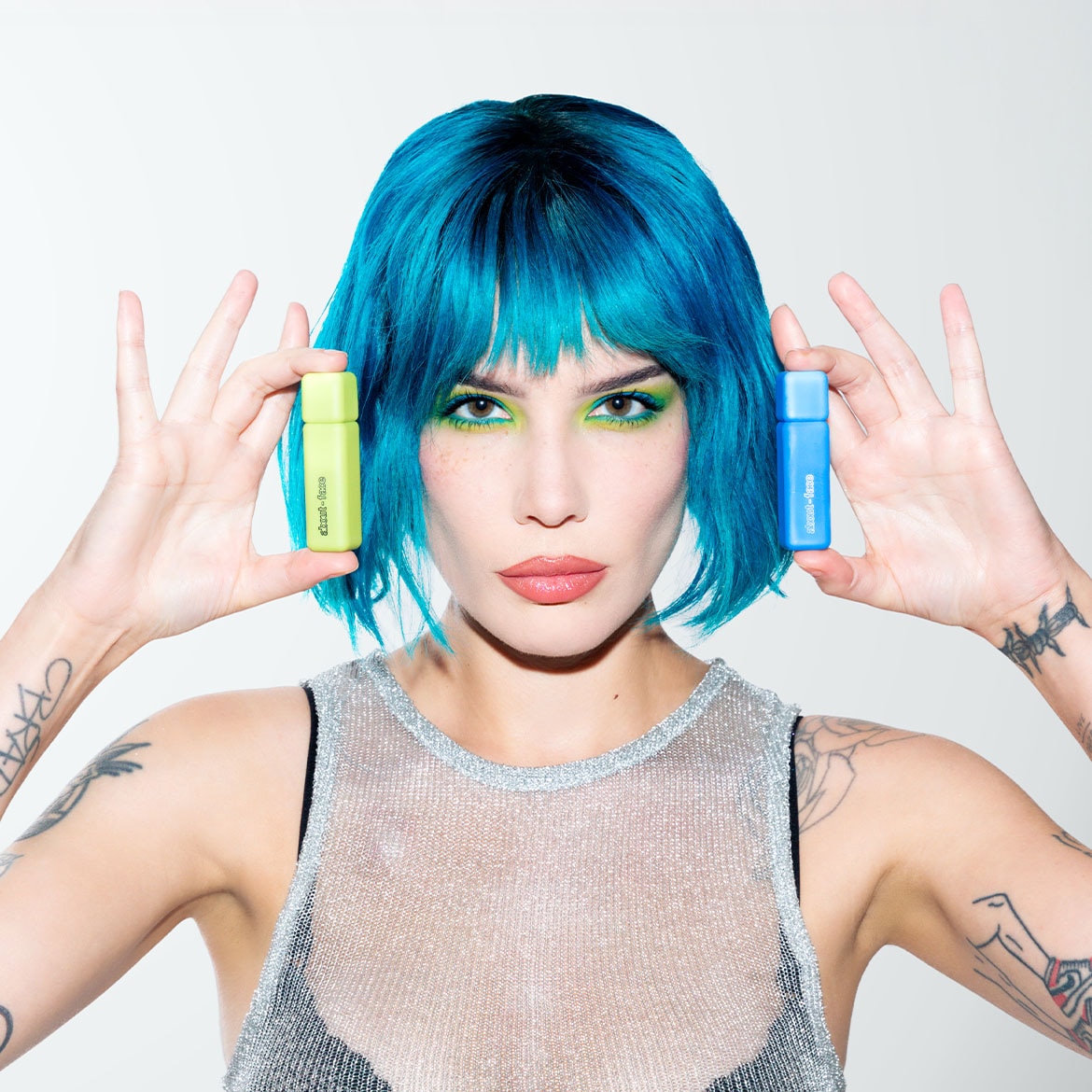 Žena s modrými vlasmi drží v každej ruke malú fľaštičku, jednu svetlozelenú a druhú modrú, pričom obe majú na sebe nápis.