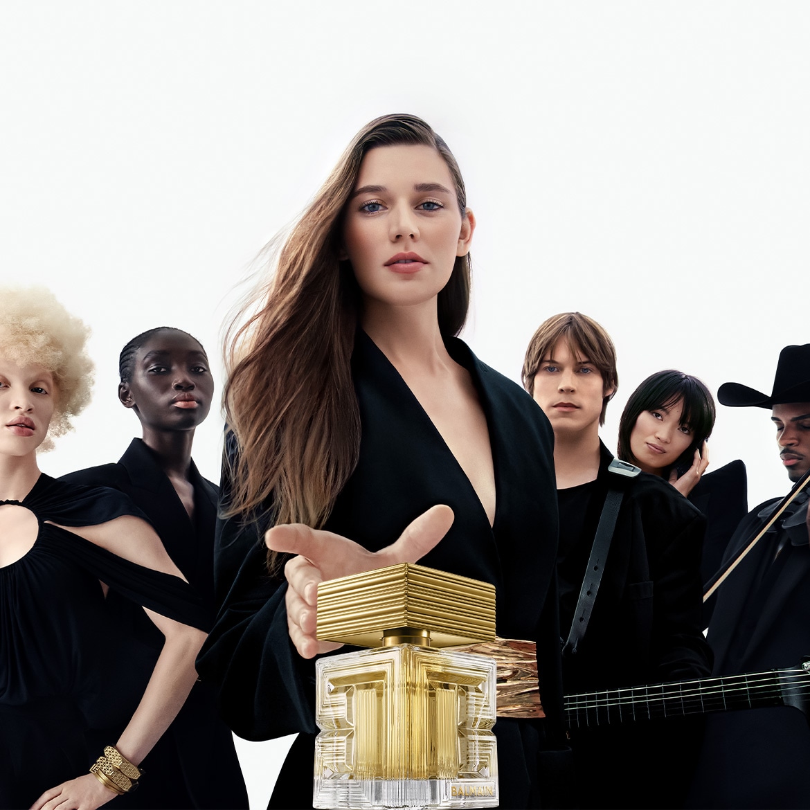 V popredí stojí flakón Balmain Paris so zlatým vrchnákom a priehľadným telom, za ktorým stojí modelka v čiernom s ďalšími modelmi a hudobníkmi v pozadí.