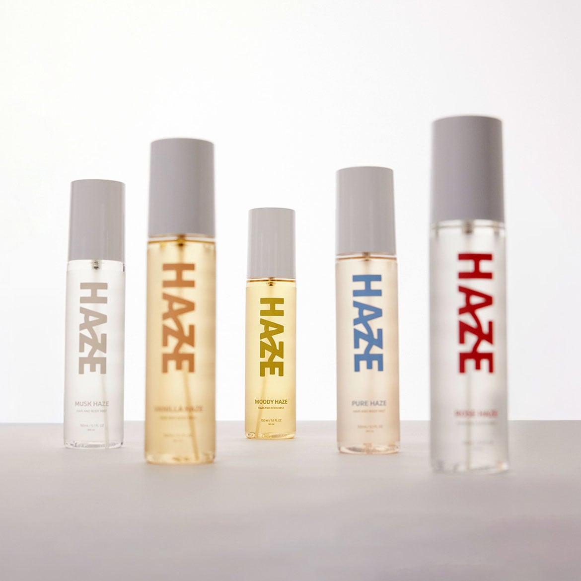 Päť fliaš parfumov Haze rôznych vôní, vrátane Musk Haze, Vanilla Haze, Woody Haze, Pure Haze a Kiss Haze, s minimalistickým dizajnom a sivými vrchnákmi.