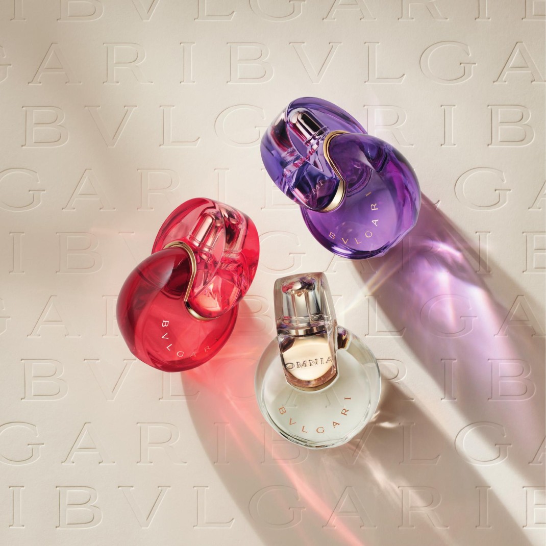 Tri parfumové flakóny Bvlgari Omnia v tvare prepojených kruhov, jeden červený, jeden fialový a jeden číry, sú usporiadané na svetlom pozadí s vyrazeným logom Bvlgari.
