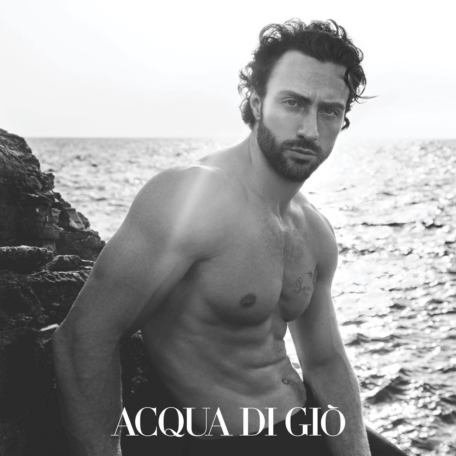 Čiernobiela fotografia muža s tmavými kučeravými vlasmi a strniskom, ktorý sedí na skalách pri mori s nápisom ACQUA DI GIÒ v dolnej časti.