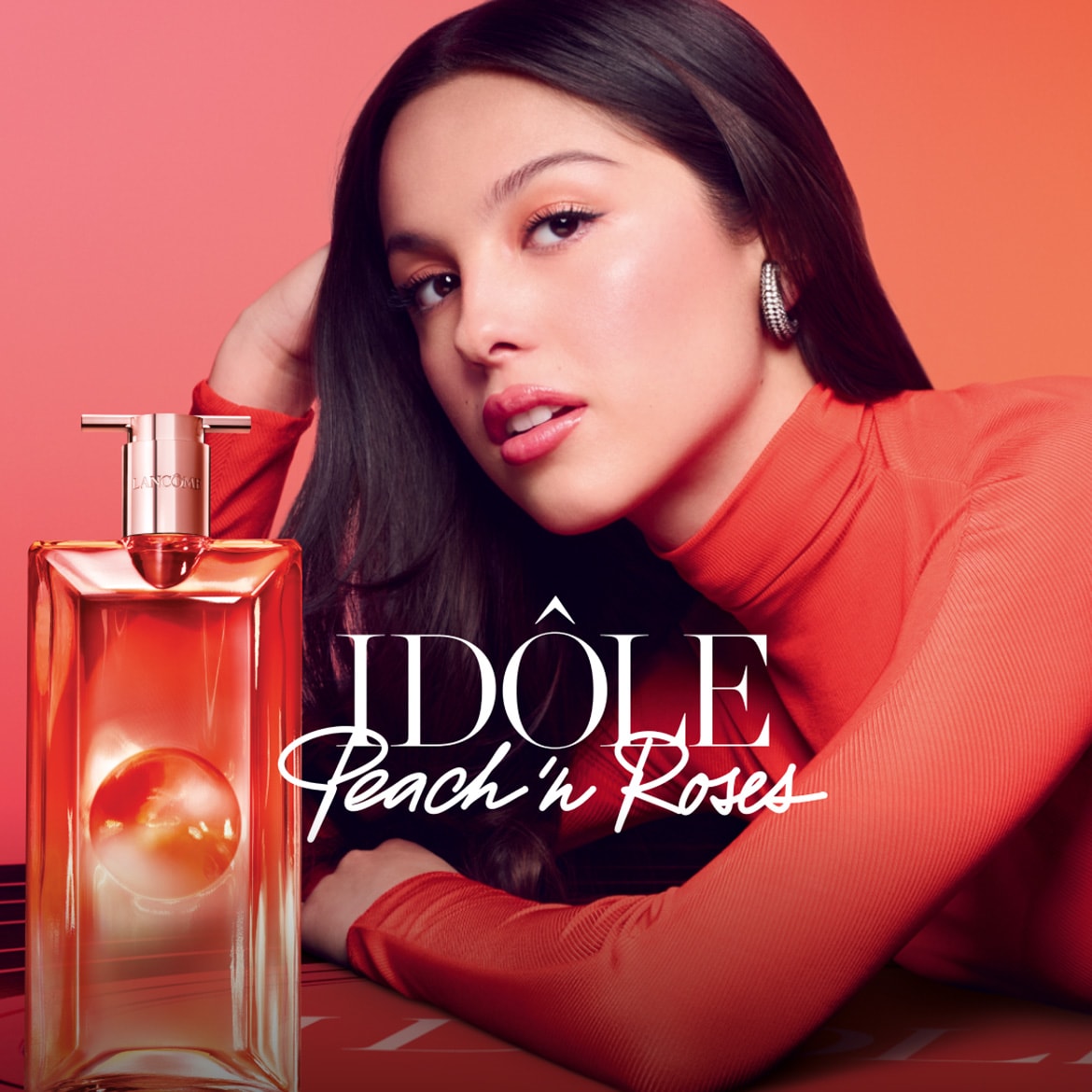 Olivia Rodrigo propaguje vôňu Idôle Peach 'n Roses od Lancôme, ktorá sa nachádza vedľa nej v priehľadnej fľaši s ružovo-zlatým uzáverom.