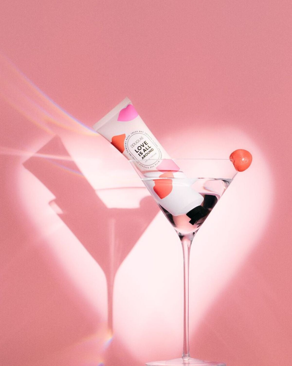 Na ružovom pozadí je koktailový pohár s ružovým ovocím na okraji, v ktorom je ponorená biela tuba s nápisom Douglas Love Is All Around.
