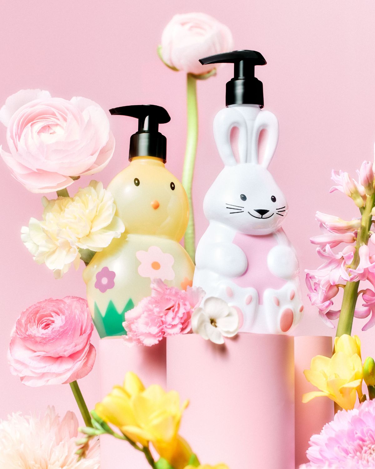 Na ružovom pozadí stoja dávkovače mydla v tvare kuriatka a zajačika, obklopené ružovými, bielymi a žltými kvetmi, ktoré vytvárajú hravú a sviatočnú atmosféru.