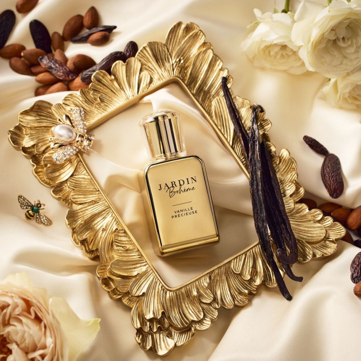 Zlatý rám s parfumom Jardin Bohème Vanille Précieuse, vanilkovými strukmi, mandľami a kvetmi na hodvábnej látke vytvára luxusnú a zmyselnú kompozíciu.