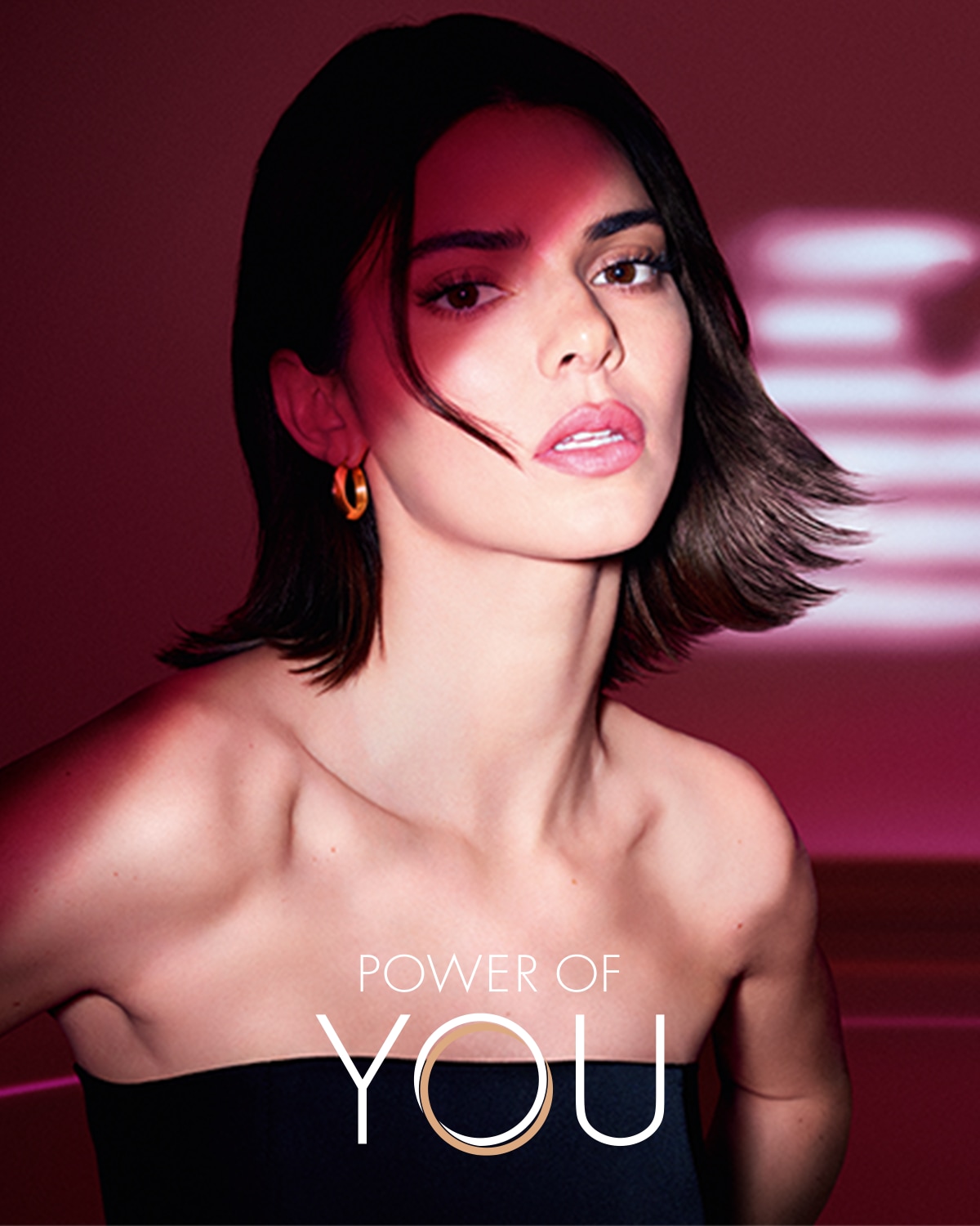 Na portréte je žena s hnedými vlasmi a ružovým pozadím, ktorá nosí čierny top bez ramienok a zlaté náušnice, s nápisom "POWER OF YOU" v spodnej časti.