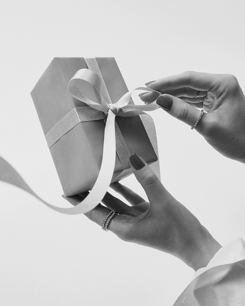 Žena s červenými nechtami rozväzuje bielu stuhu na sivom darčekovom balení, na prstoch má prstene.